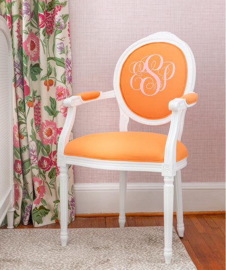 White Louis style arm chair in children's nursery with light pink walls.  Custom upholstered chair has orange fabric and chair back has a pink embroidered monogram.