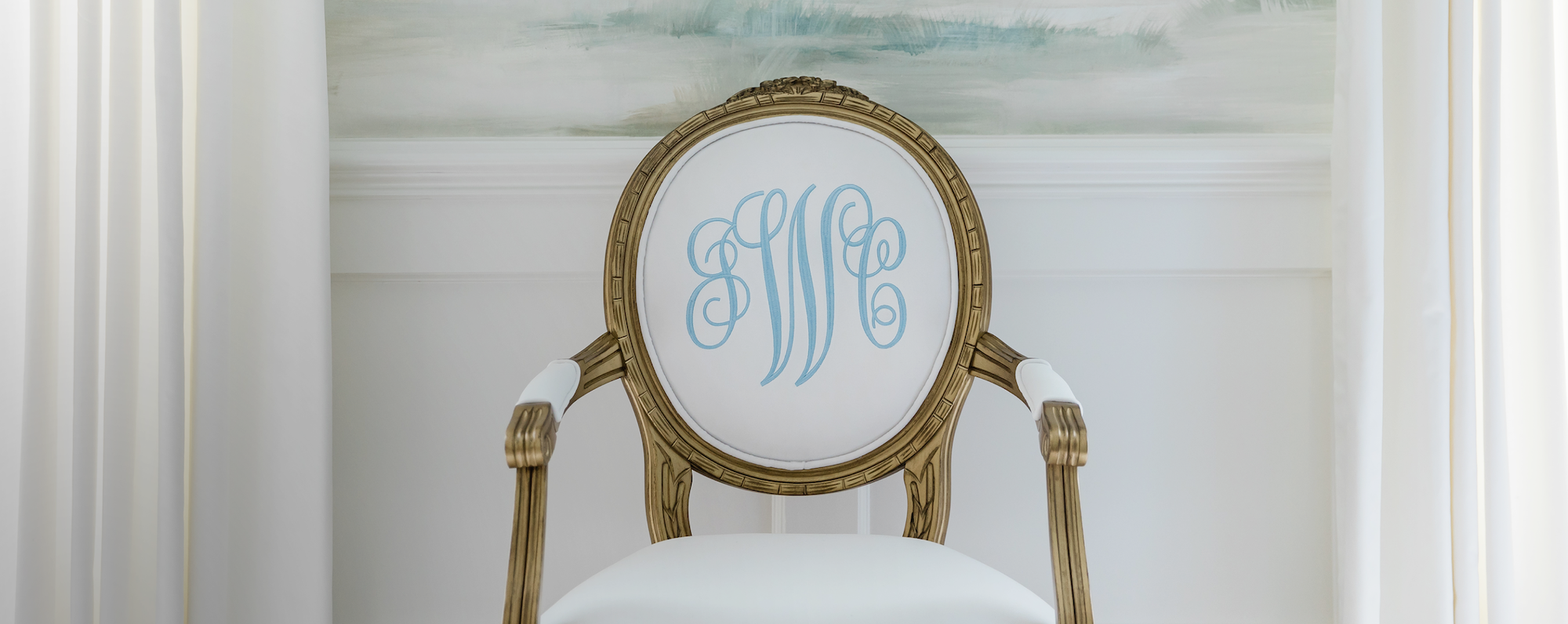 French gold King Louis XVI Oval back Arm Chair. Custom dining chair personalized with light blue embroidered monogram on white velvet fabric. Monogrammed Chair is sitting in front of a wallpapered Dining Room. Embroidered custom and monogrammed chair.