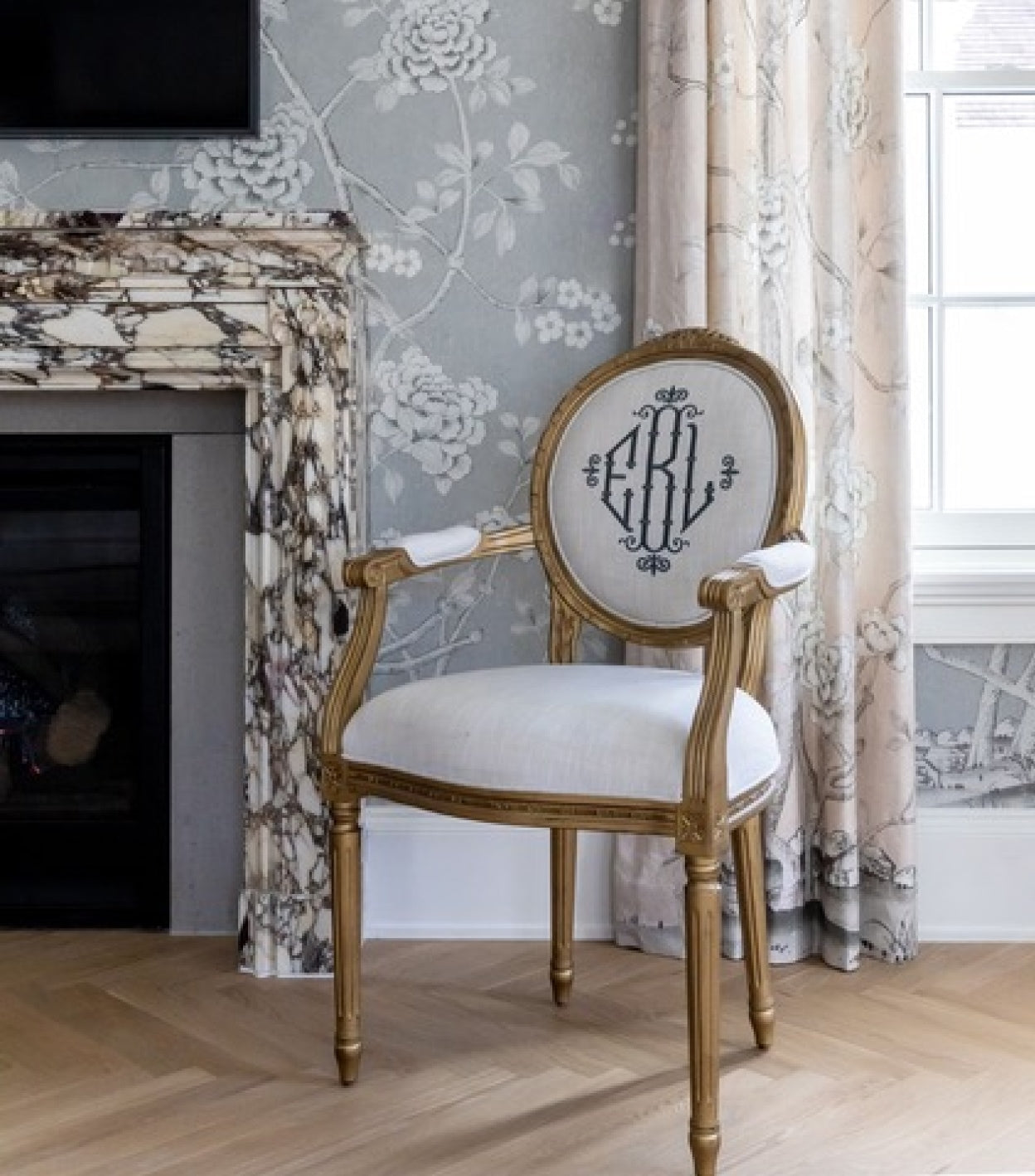 Gold King Louis XVI Oval Back Arm Chair with neutral linen fabric and grey embroidered monogram. Custom upholstered chair is sitting beside a bedroom fireplace with marble surround and floral wallpaper designed by The Fox Group.