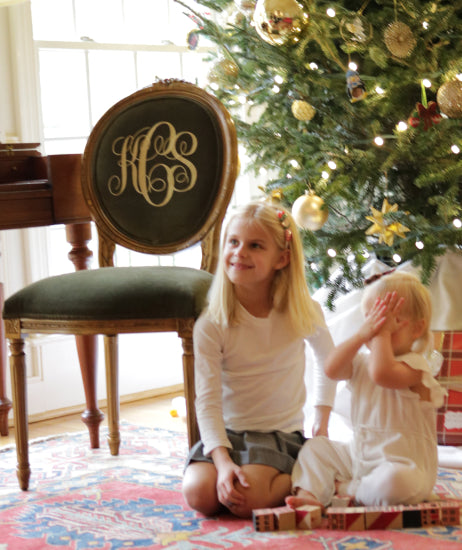 Two little girls sitting on a rug in front of a Christmas tree. Beside them is a custom Louis style chair. Chair has a gold finish, dark green velvet upholstery and personalized chair back with an embroidered monogram in gold thread.
