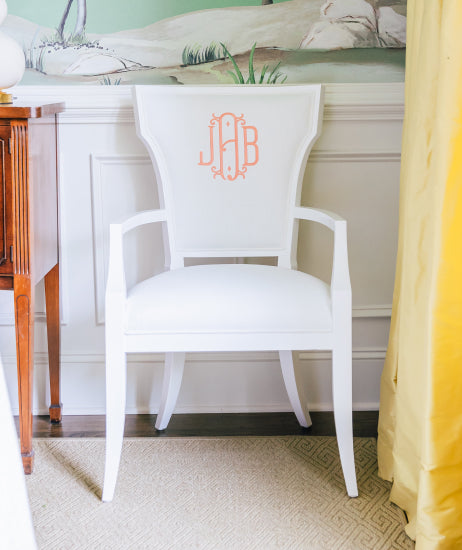 All white flare back arm chair sitting against a wallpapered Dining Room wall with white wainscoting. The inside of the custom upholstered chair back features  a coral, three-initial monogram.