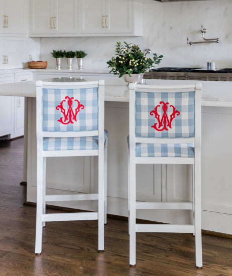 Back side of two blue and white monogrammed barstools sitting at a white kitchen island with marble countertop. White wood upholstered barstools have a light blue and white check fabric and red embroidered monograms on the barstool backs.