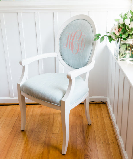A sweet children's chair in the corner of a bedroom. White wood arm chair has a round back, sky blue linen fabric and light pink monogram. Custom made.
