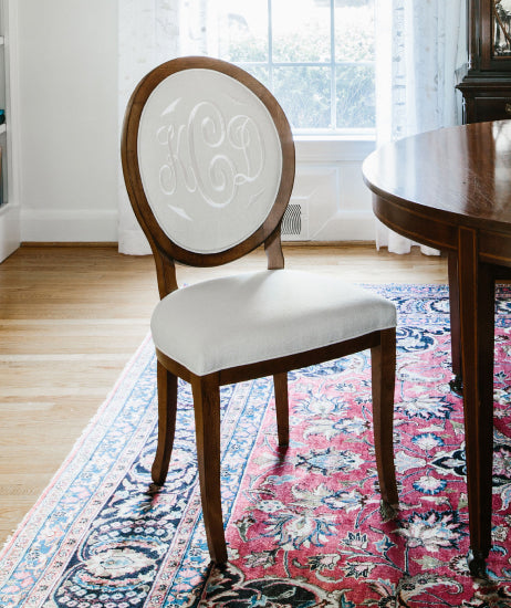 Brown wood side chair at a dining room table. Custom upholstered chair features white linen fabric and matching white embroidered monogram with burnished walnut wood finish. Oriental style rug under the table. Traditional dining room chair.