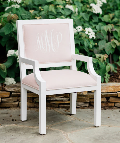 White custom kids chair with light pink velvet fabric sitting in front of a flower bed. Personalized chair has a white embroidered monogram on the inside back. Sweet nursery chair and personalized gift for children.