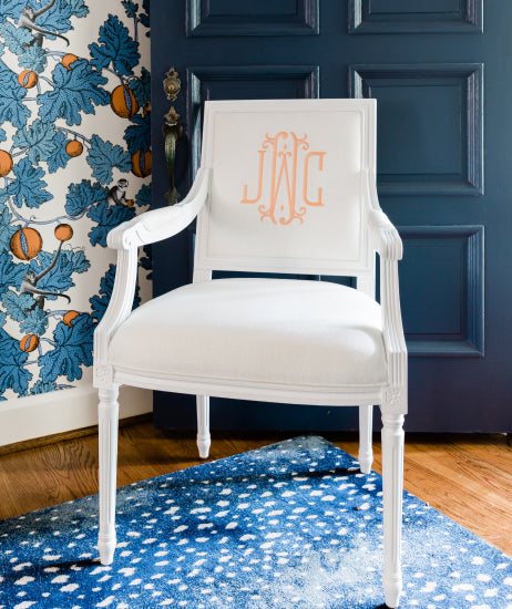 All white square back Louis XVI arm chair sitting in a foyer in front of a navy blue door, blue and white spotted rug and wallpaper with blue leaves and oranges. Custom upholstered chair features a personalized chair back with a tangerine three-initial embroidered monogram. Beautiful modern monogrammed chair and room.