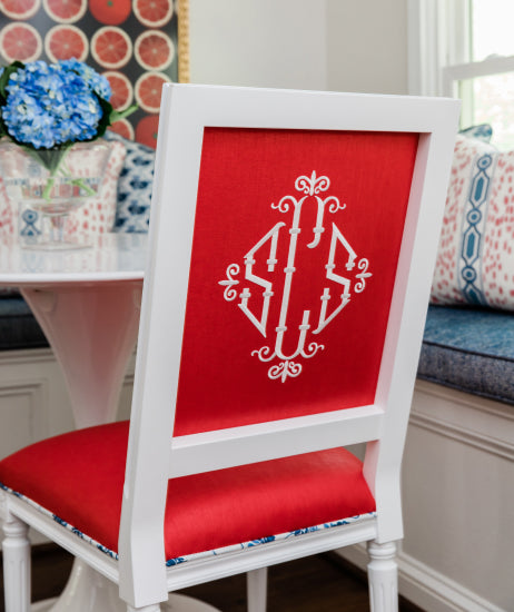 The back of a white square back dining chair with red linen fabric and a large, white three-initial monogram. Custom upholstered monogrammed dining chair sits at a kitchen table in a breakfast nook with red, white and blue accents.