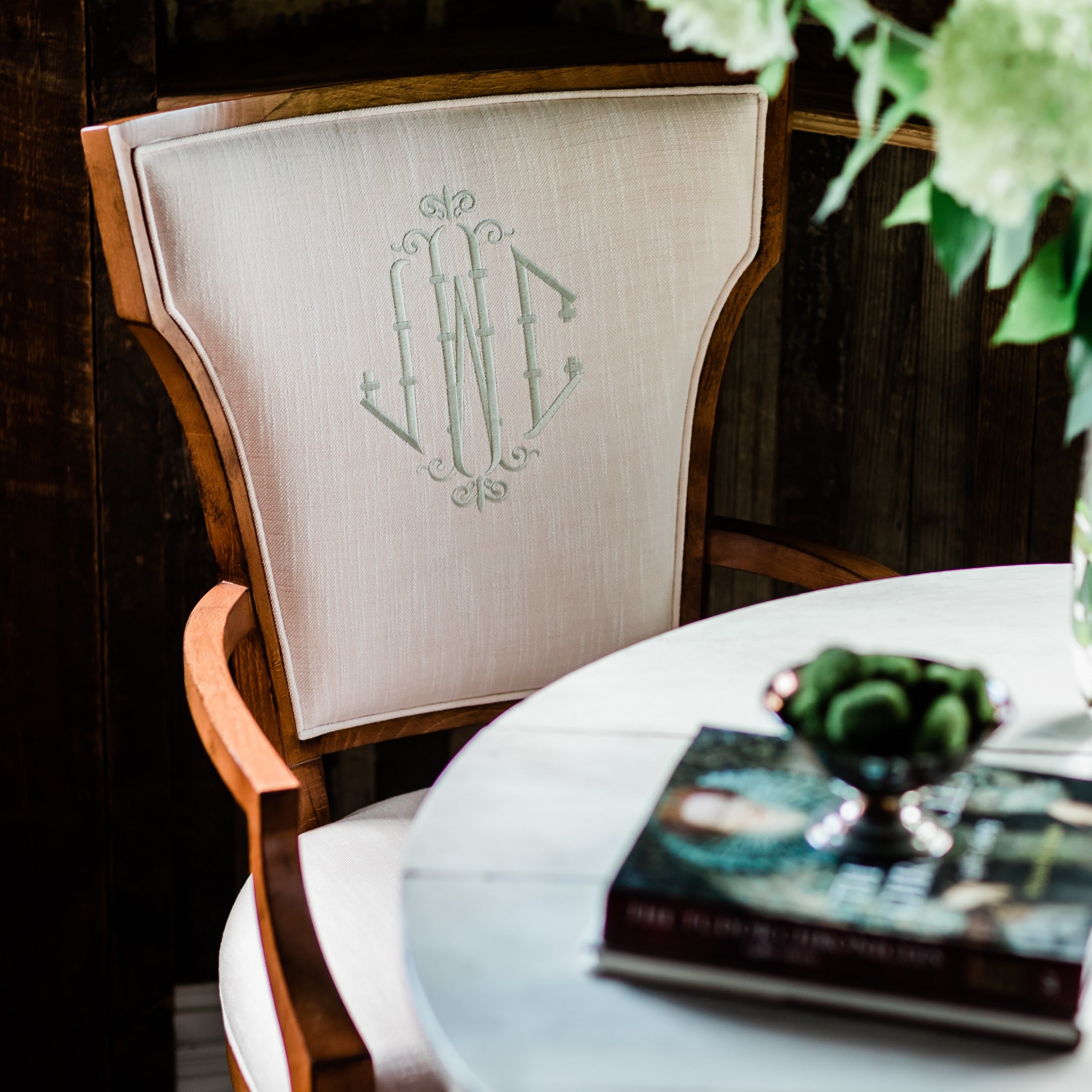 Moody burnished walnut arm chair at a white breakfast table. Custom chair personalized with light tan embroidered monogram. Dining chair.