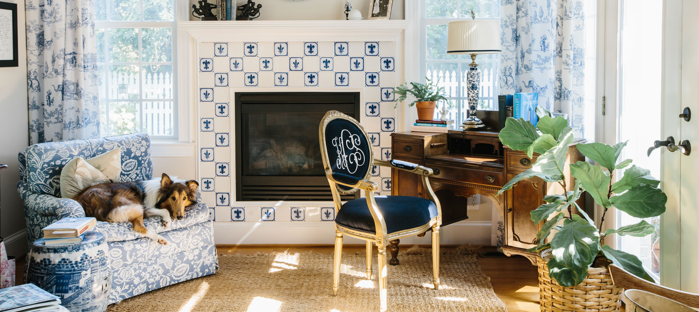 Gold Louis style arm chair with navy blue velvet and white embroidered monogrammed chair back. Custom desk chair in a blue and white sunroom with fireplace and dog sitting on nearby upholstered chair.