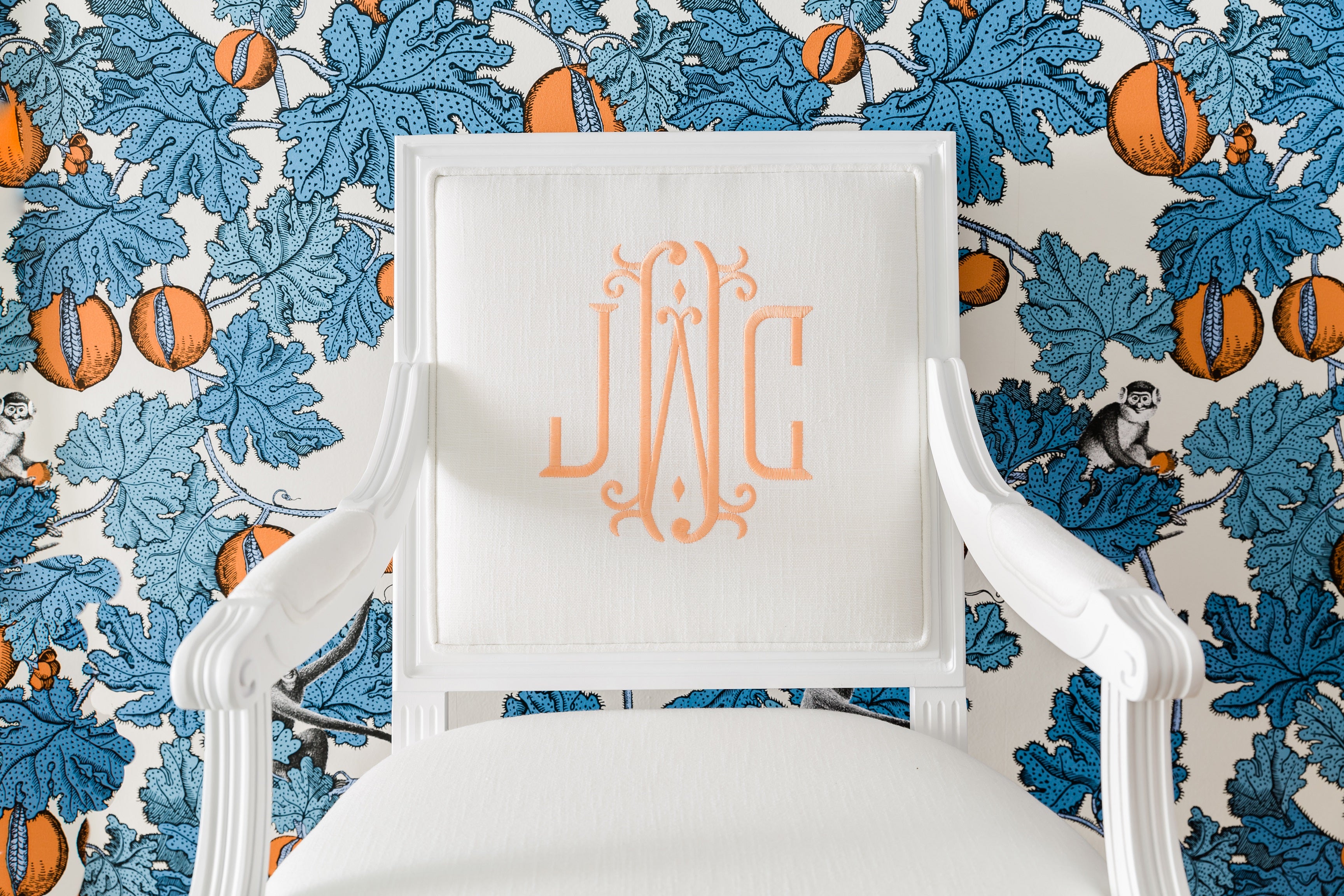 White custom arm chair with white linen fabric and light orange embroidered monogram. Monogrammed chair sits in front of a statement blue leaf wallpaper with orange tangerines.