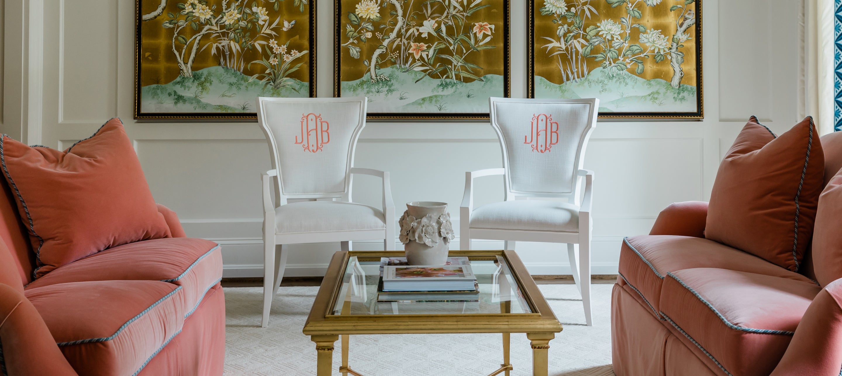 Two white custom upholstered arm chairs with flared backs sit in a formal living room. Chair fronts are personalized with embroidered monograms in a coral thread.