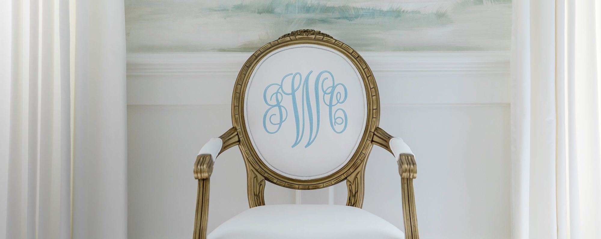 French Gold King Louis XVI Oval Back Arm Chair. Personalized Chair with blue script monogram on white velvet fabric. Monogrammed Chair is sitting in front of a wallpapered Dining Room. Embroidered and monogrammed chair.